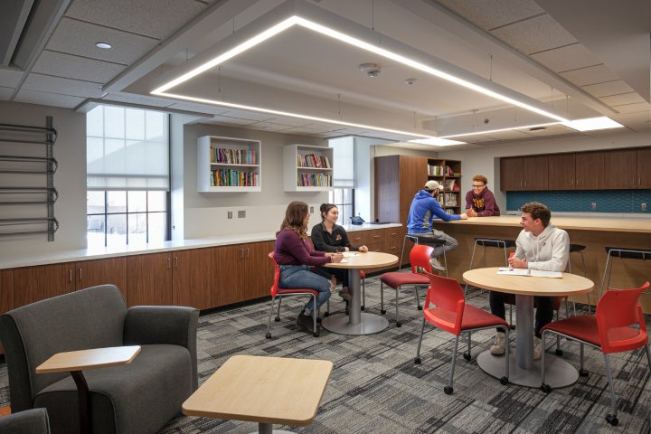 A modern study lounge with wooden cabinetry, bookshelves, and small round tables. Several students sit and talk, while two others stand at a counter in the background. The space features comfortable seating and a well-lit atmosphere